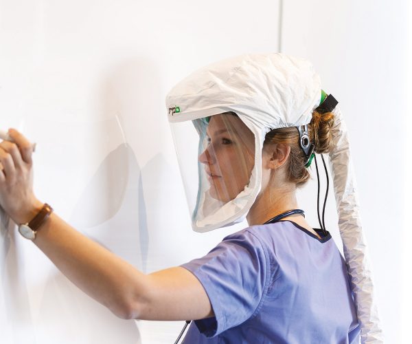 A healthcare worker wearing a respiratory protective hood with a clear visor and a white air supply hose