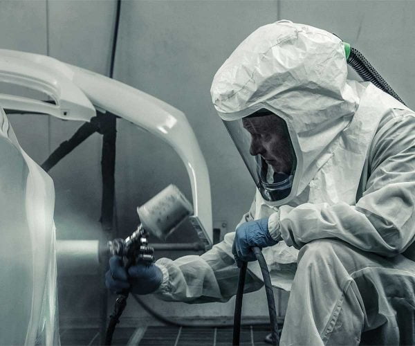 Person in a protective RPB safety hood and suit, using a spray gun in an industrial setting.