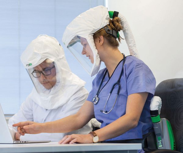 Two healthcare professionals in respiratory protective equipment and protective gear work together at a laptop.