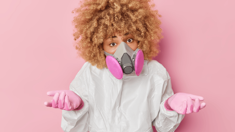 Person in a white protective suit and pink gloves, shrugging while wearing a GVS Malaysia respirator with pink filters, against a pink background, conveying confusion or uncertainty about safety protocols.