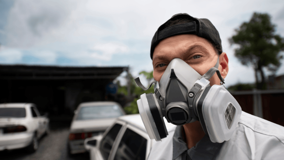 Close-up of a man wearing a GVS Malaysia respirator, with a black cap and a light shirt, confidently smiling in an outdoor setting with cars and trees in the background.